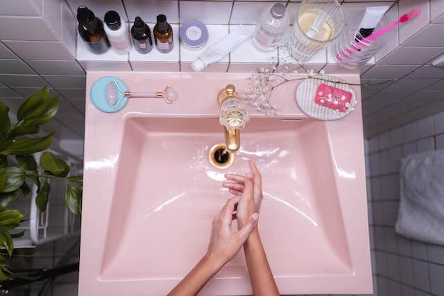 Uma pia cor rosa, muito limpa e organizada, com vários produtos de higiene pessoal, com uma pessoa lavando as mãos: Limpa Fossa Planalto.